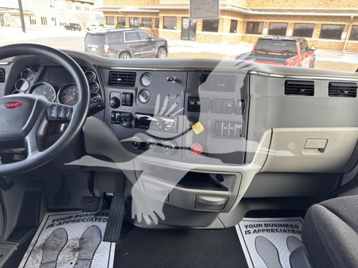 Peterbilt 579 truck interior dashboard with steering wheel on left, center console, and driver-side footwell; visible text includes 'Peterbilt' on steering wheel emblem, various control labels on the dashboard, and floor mats with text (not