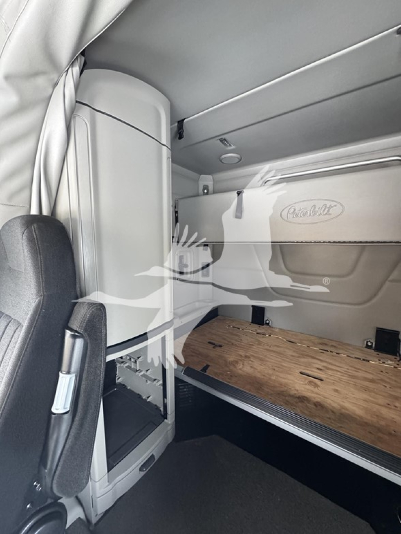 Peterbilt 579 interior sleeper cab with wooden bed platform, side storage panel, and logo reading Peterbilt. Cabin area shows side wall panels and overhead compartments.