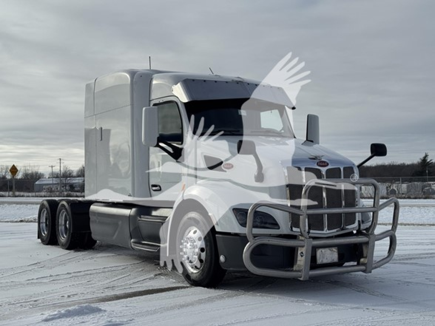 Peterbilt 579 tractor in snowy lot, silver cab, chrome bumper guard, dual rear wheels, manufacturer badges visible on grille, no visible text legible other than truck design.