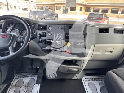 Peterbilt 579 truck interior dashboard with steering wheel on left, center console, and driver-side footwell; visible text includes 'Peterbilt' on steering wheel emblem, various control labels on the dashboard, and floor mats with text (not