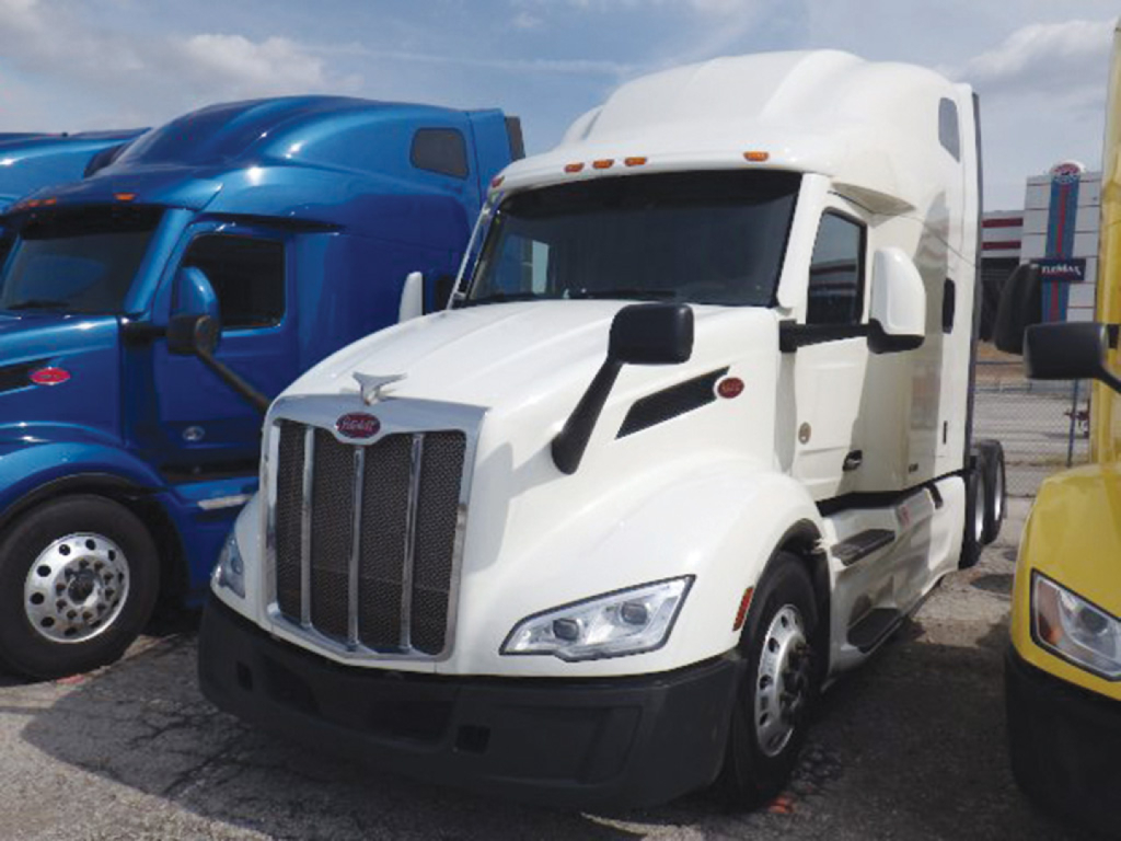 Peterbilt 579 EPIQ conventional sleeper truck, white cab, front grille with vertical bars, parked among blue and yellow trucks.