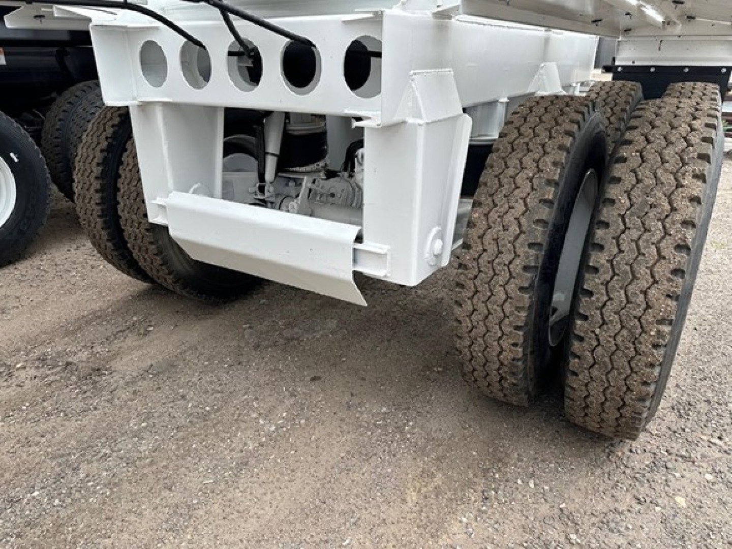 Rear view of a white dump trailer chassis showing dual-axle tires with textured treads and a heavy steel frame, with visible suspension and undercarriage components.