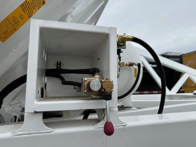L ехor: Valve assembly and hoses on a white dump trailer, showing a small brass valve with a white knob inside a rectangular white box, and a red drop/plug below; hydraulic line connections visible.
