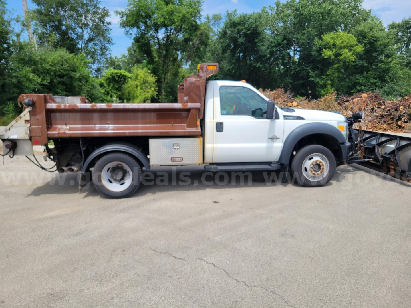 Ford F-550 dump truck side view showing white cab and brown dump bed with utility compartments, plow equipment visible in the background.