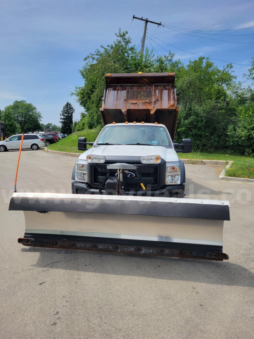 Front view of a 2012 Ford F-550 with a large snow plow attached, truck in a parking lot, dump bed raised in background.