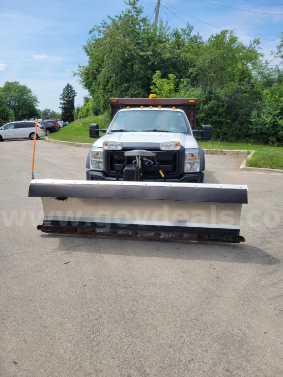 Front view of a white 2012 Ford F-550 with snow plow attachment, headlights, grille, and towing hitch visible.
