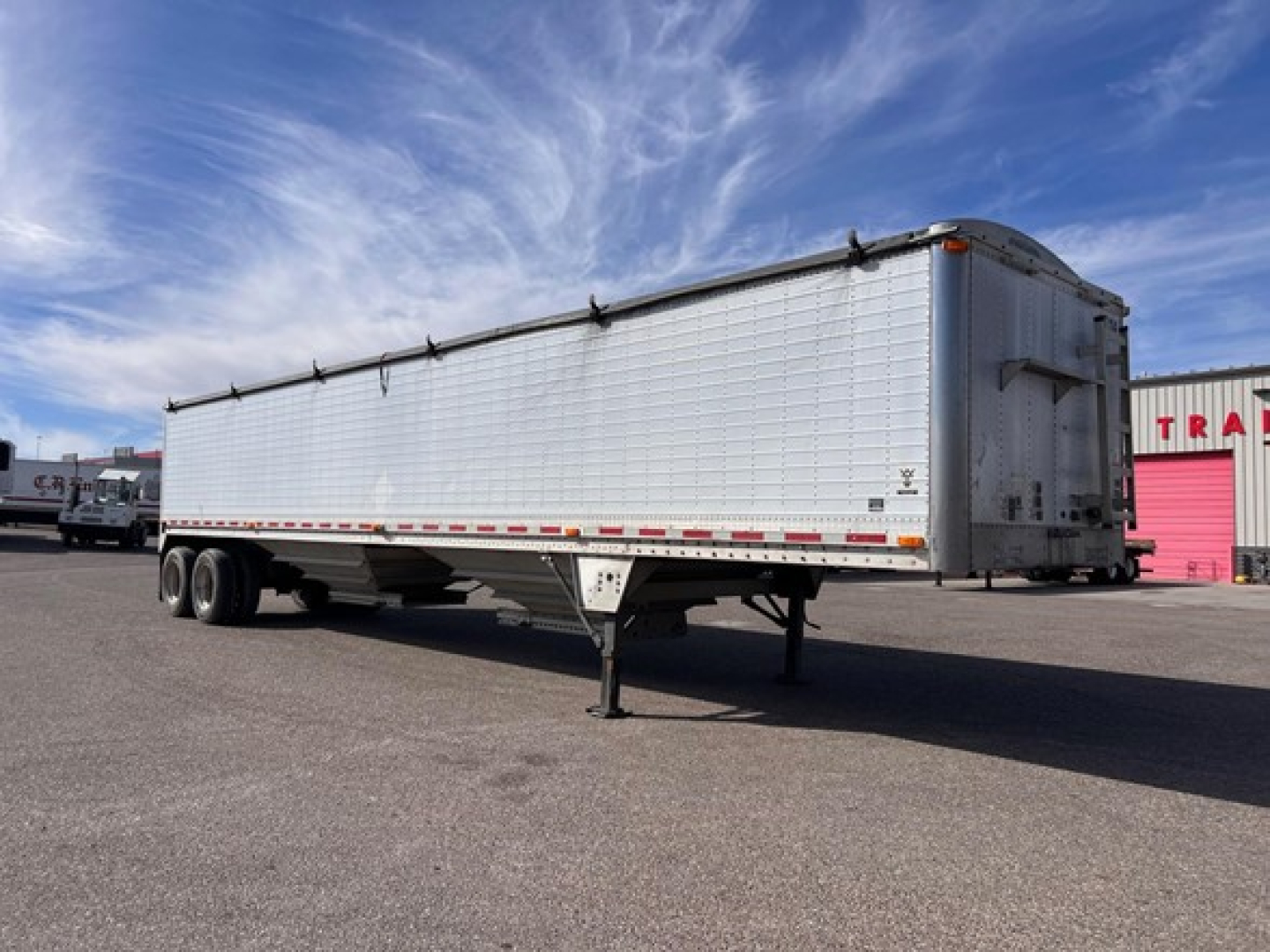 Trailer side view showing a long silver Wilson hopper trailer with a raised tarp, undercarriage with dual rear axles, and a front landing gear with a white wheel.