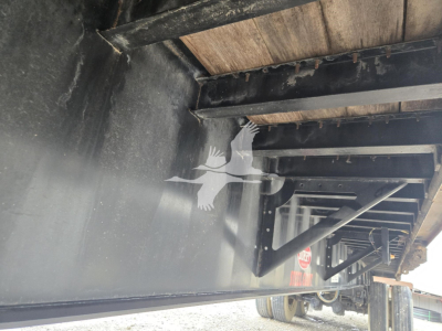 Flatbed trailer undercarriage with structural beams and wheel area visible; a faint bird logo overlain due to watermark; no legible model text readable in the image.