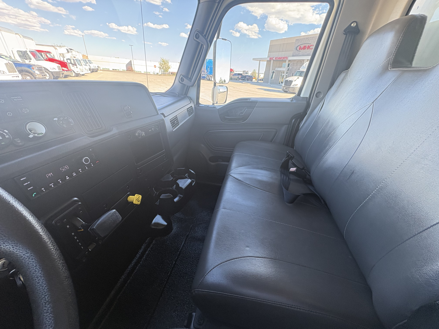 Truck cab interior with dashboard controls and front bench seat of a 2021 International MV607, steering wheel visible on left, manual gear shifter, large side window views.