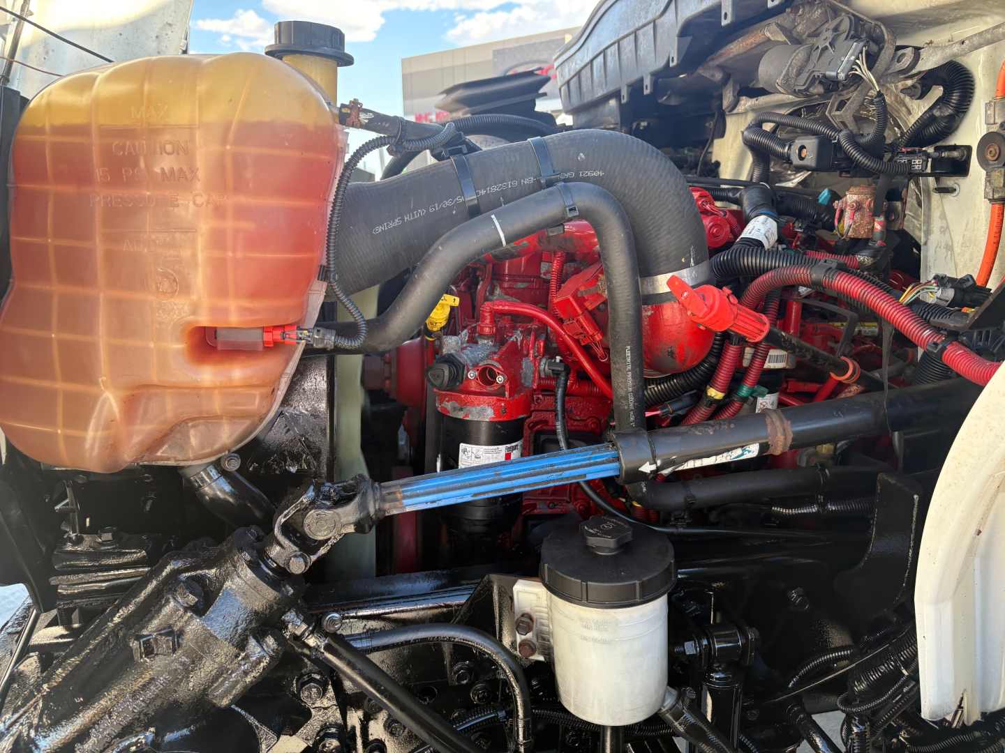 Truck engine bay of a 2021 International MV607 showing coolant reservoir, red engine components, hoses, and fluid reservoirs.
