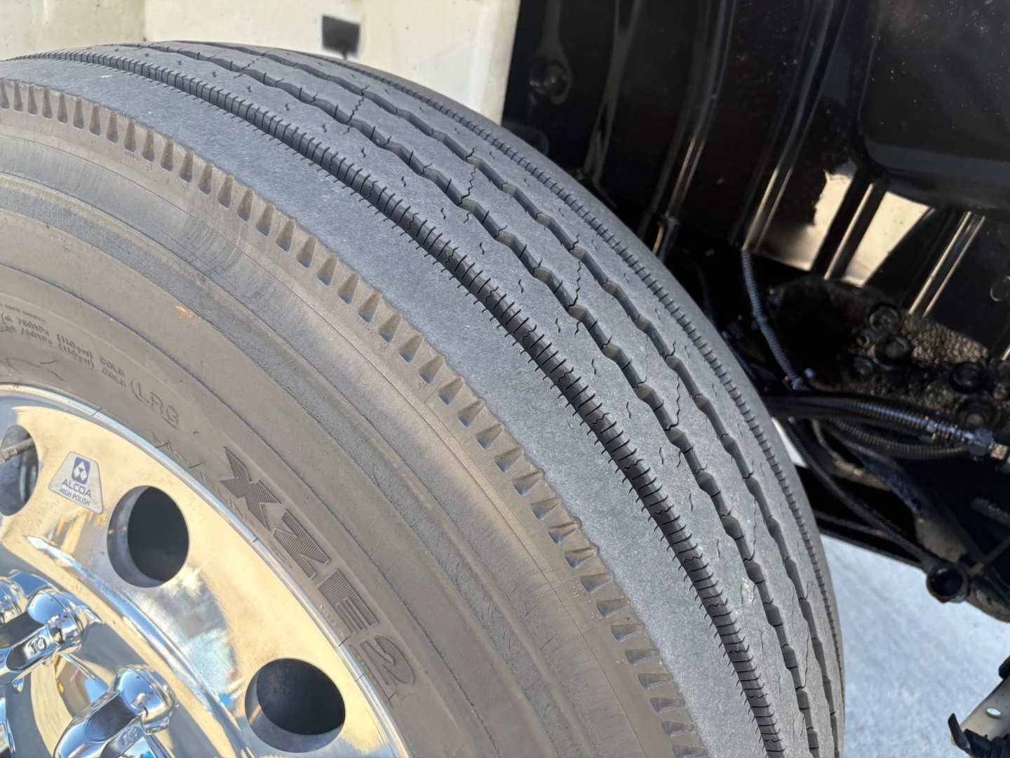 Truck wheel and tire with visible tread and hub, part of International MV607 front suspension; rim shows stamped numbers and lug nuts; components in view include tire sidewall details