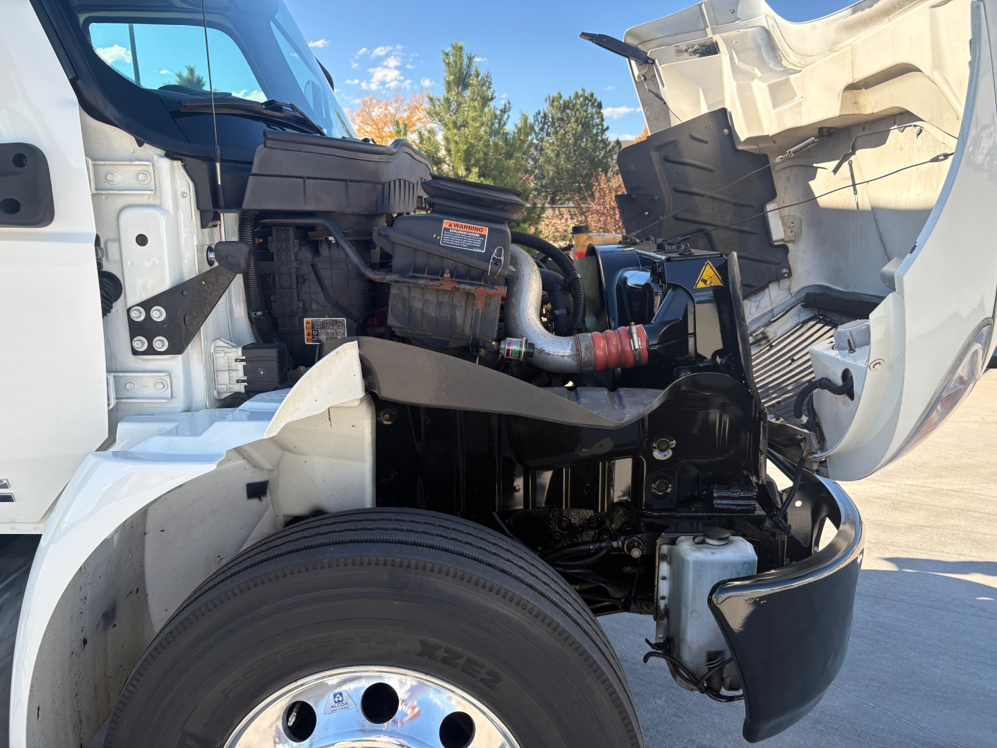 Truck engine bay with open hood showing air intake, hoses, and battery; front wheel and suspension visible; components labeled but no legible text clearly readable.