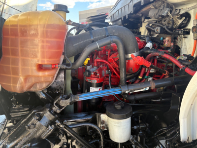 Truck engine bay of a 2021 International MV607 showing coolant reservoir, red engine components, hoses, and fluid reservoirs.