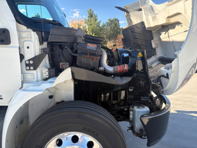 Truck engine bay with open hood showing air intake, hoses, and battery; front wheel and suspension visible; components labeled but no legible text clearly readable.