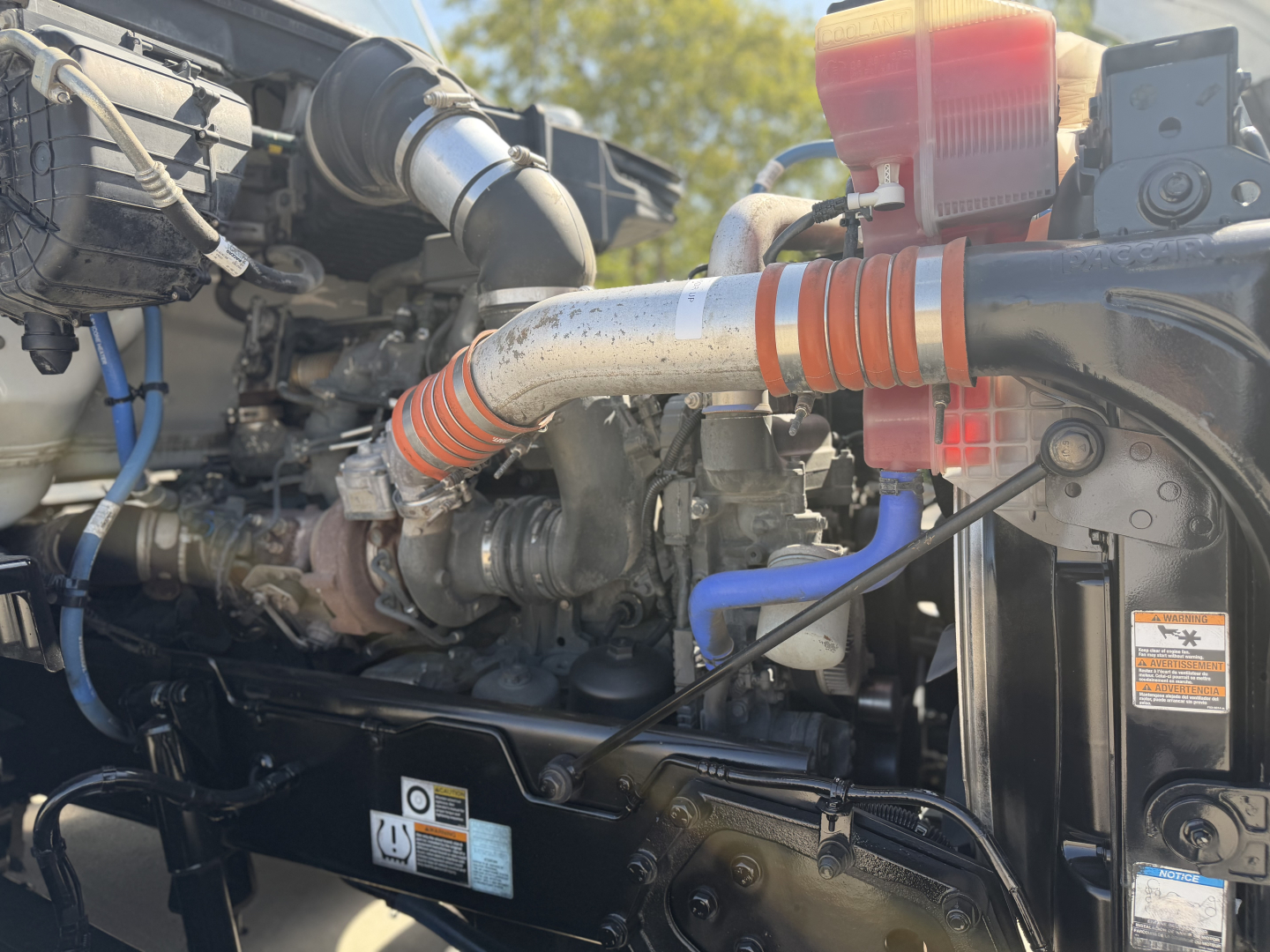 Kenworth truck engine bay with large orange coolant hose, silver intercooler pipe, red coolant reservoir, black radiator, blue coolant line, and various hoses. Visible labels and stickers on components.