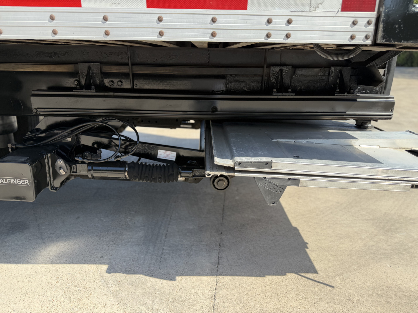 Kenworth T680 rear underbelly showing hydraulic system and cargo box with loading step and label areas.