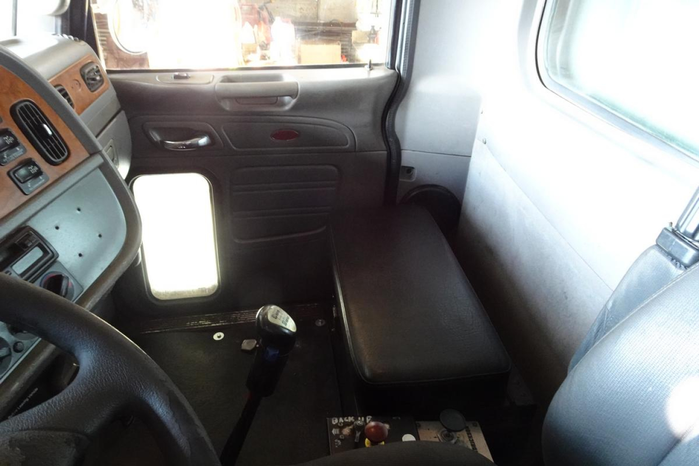 Peterbilt truck cab interior shows seating area with bench-style passenger seat, dashboard controls, gear shifter, and window view.