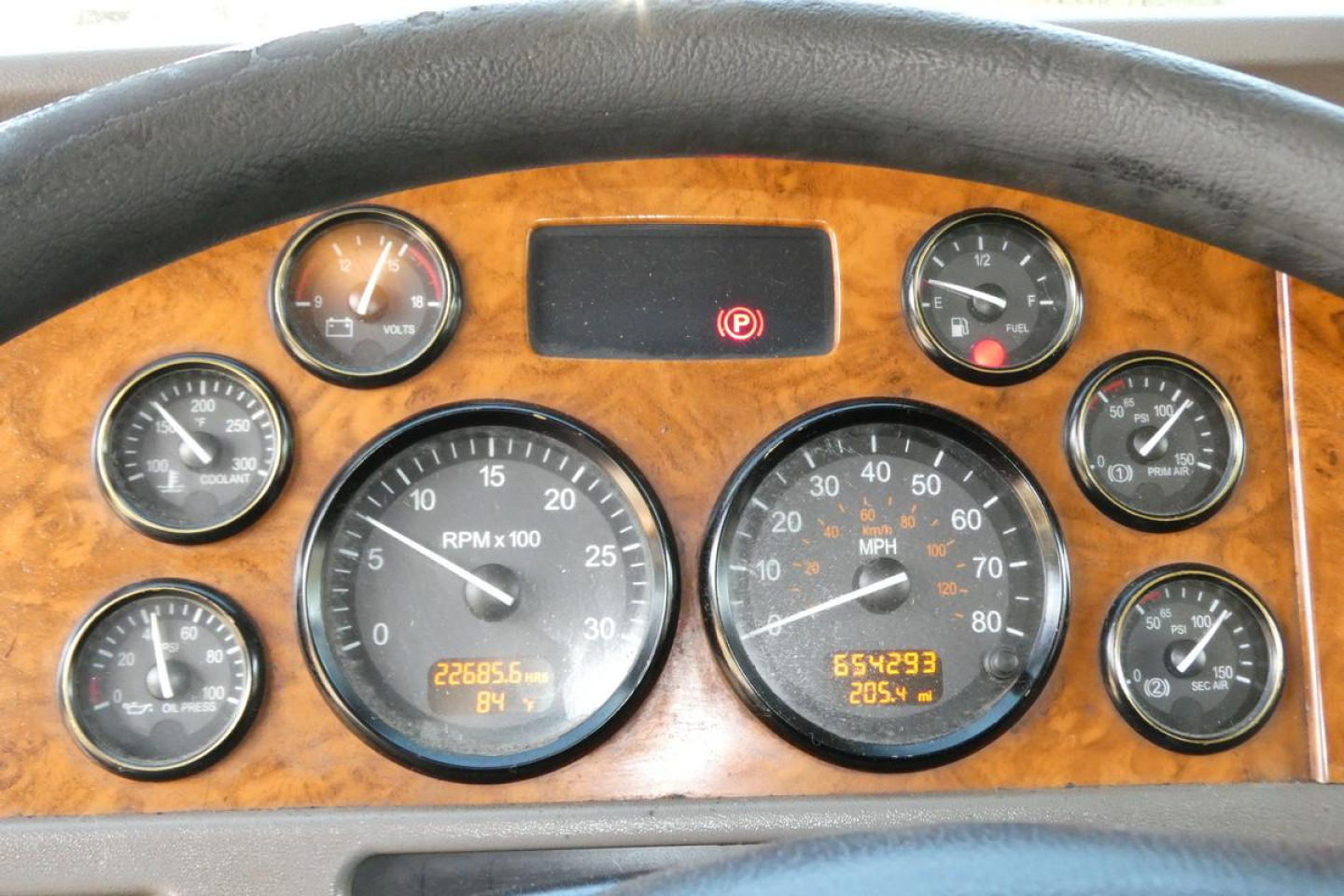 Truck dash cluster with woodgrain panel showing multiple gauges; central RPM gauge, highway speedometer, and assorted round gauges; small digital readouts at bottom; steering wheel at top edge.