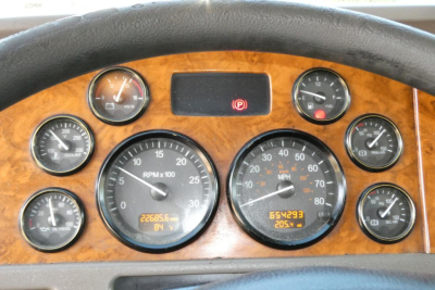 Truck dash cluster with woodgrain panel showing multiple gauges; central RPM gauge, highway speedometer, and assorted round gauges; small digital readouts at bottom; steering wheel at top edge.