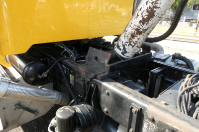Peterbilt truck engine bay area showing yellow body, exhaust pipe, and hydraulic lines with various black metal components.