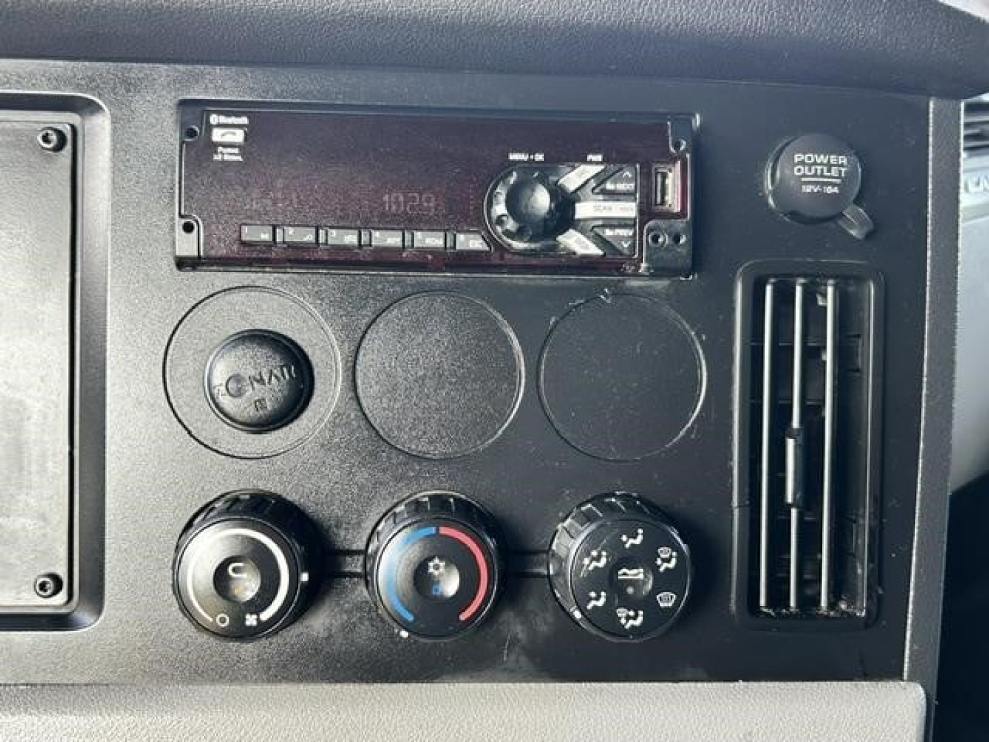 Dashboard panel with car stereo/head unit and climate controls. Visible text includes 'POWER OUTLET' on a circular cap, and parts of the stereo display and buttons.