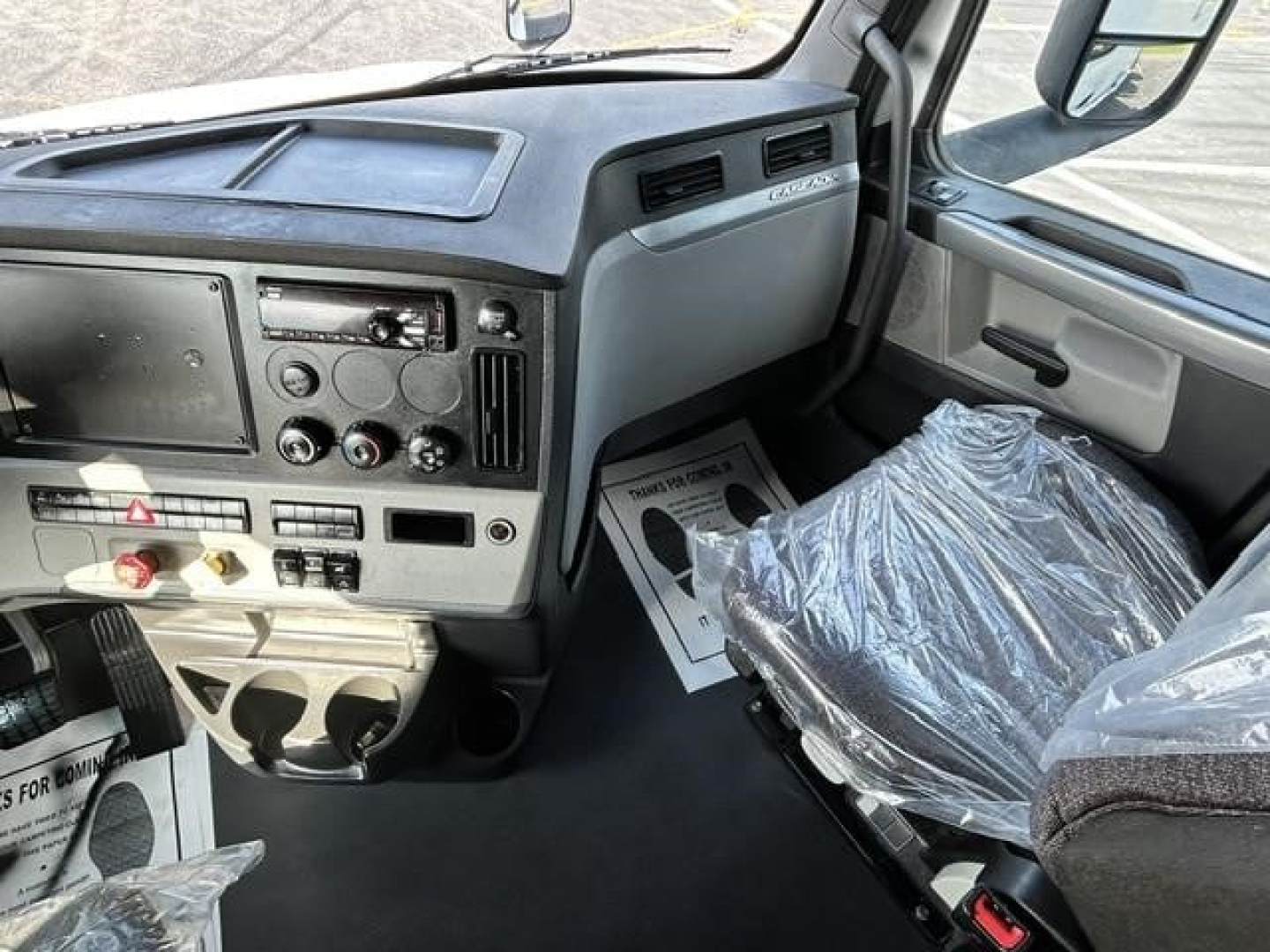 Cab interior of a 2021 Freightliner Cascadia 126 sleeper cab showing dashboard with radio, climate controls, cup holders, and seat wrapped in plastic.