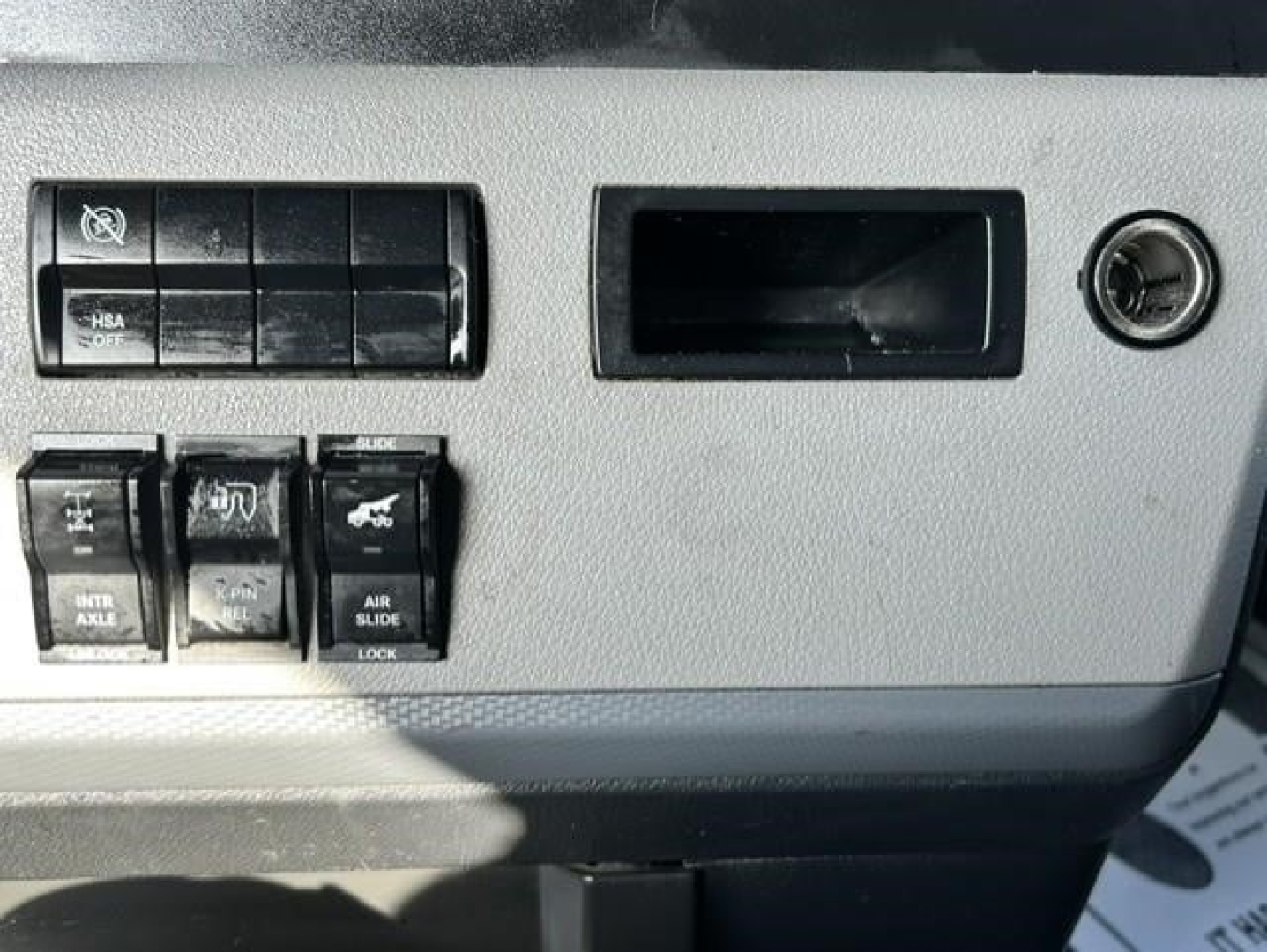 Freightliner Cascadia dash panel showing switches and blank slot; ignition barrel to right; multiple rocker switches labeled INTR AXLE, SLIDE, AIR SLIDE, and other controls.