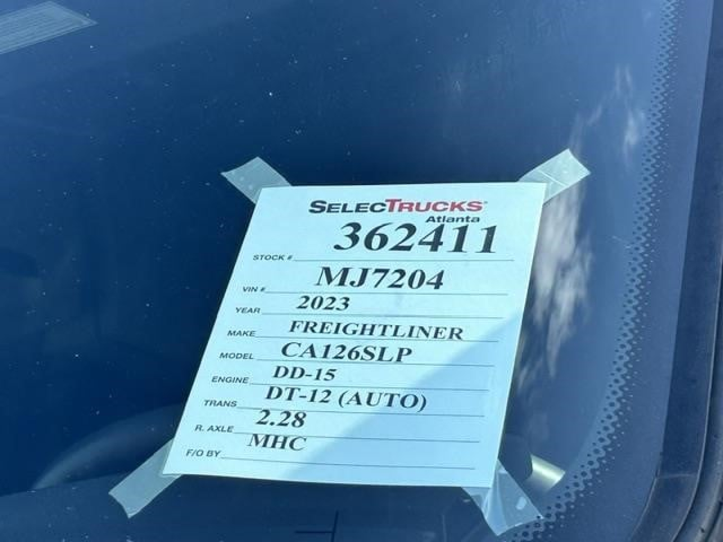 Vehicle tag on windshield with stock number 362411, label shows 2023 Freightliner model CA126SLP, engine DD-15, DT-12 (AUTO), R. AXLE 2.28, VIN MJ7204, etc.