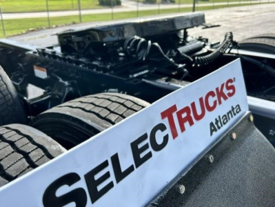 Truck chassis with heavy-duty frame and wheel, Freightliner Cascadia sleeper cab undercarriage visible, Select Trucks Atl logo panel in foreground.