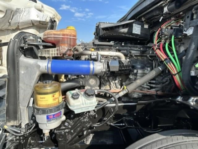 Engine bay of a Freightliner Cascadia showing blue silicone hose, coolant reservoir, power steering fluid reservoir, exposed wiring, and air intake near the fan.