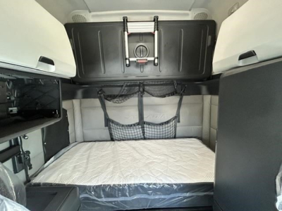 Interior sleeper cab cargo area of a 2021 Freightliner Cascadia 126 showing a bed with white mattress, overhead storage compartment, and a storage net on the wall.