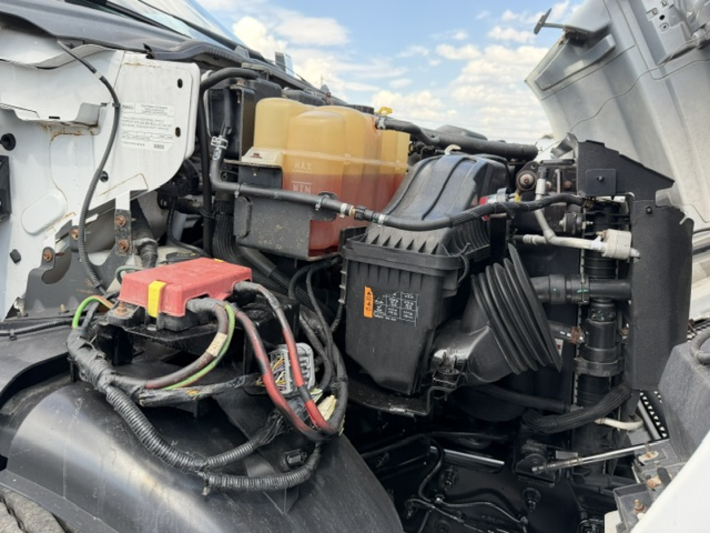 Ford F-750SD engine bay showing coolant reservoir, air filter housing, battery, wiring, and assorted hoses with engine components visible.