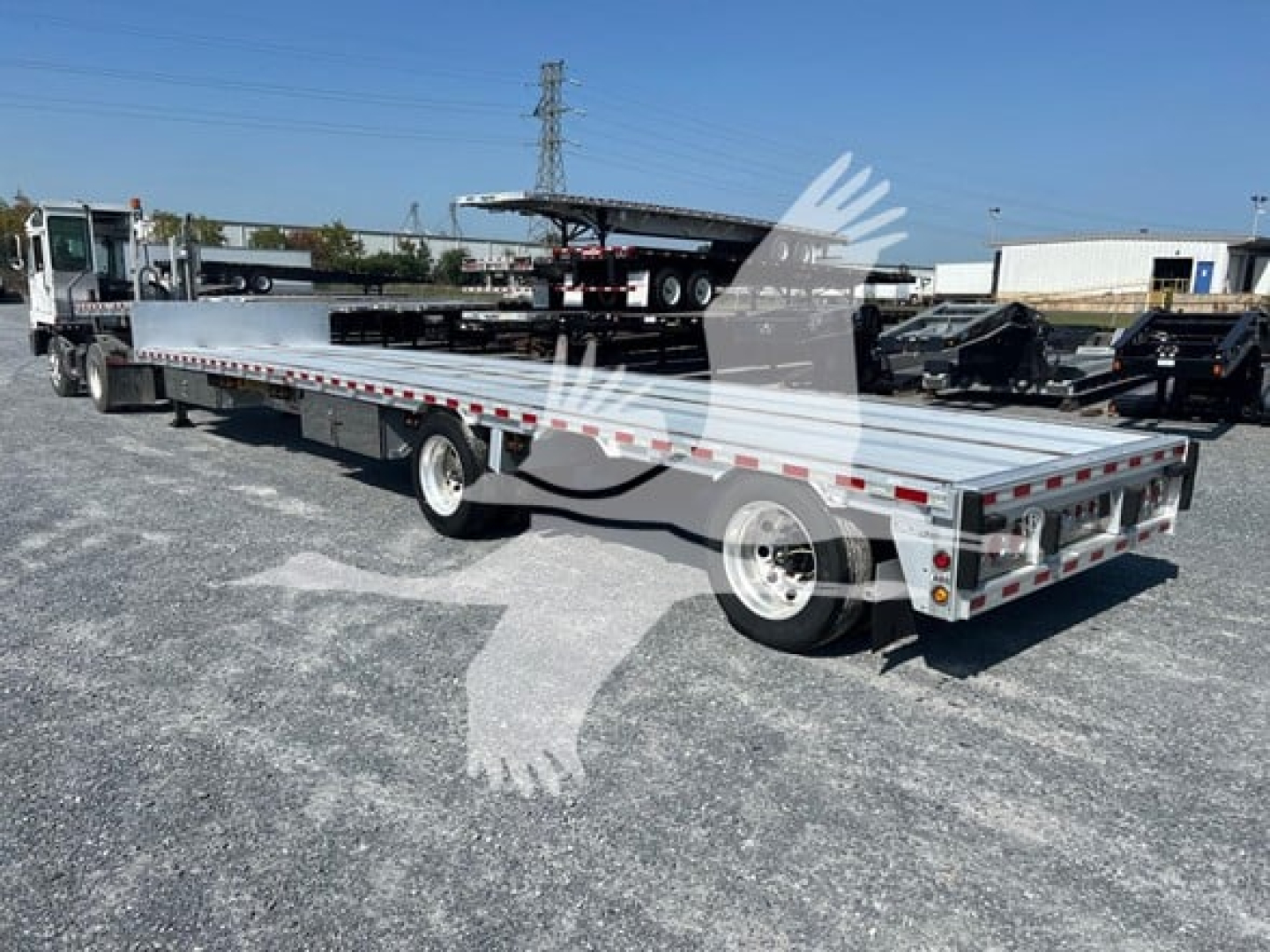 Flatbed trailer with extended drop deck showing a long aluminum platform, dual rear axles, and safety rails. Vehicle outline watermark covers center. Various equipment and other trailers visible in background.
