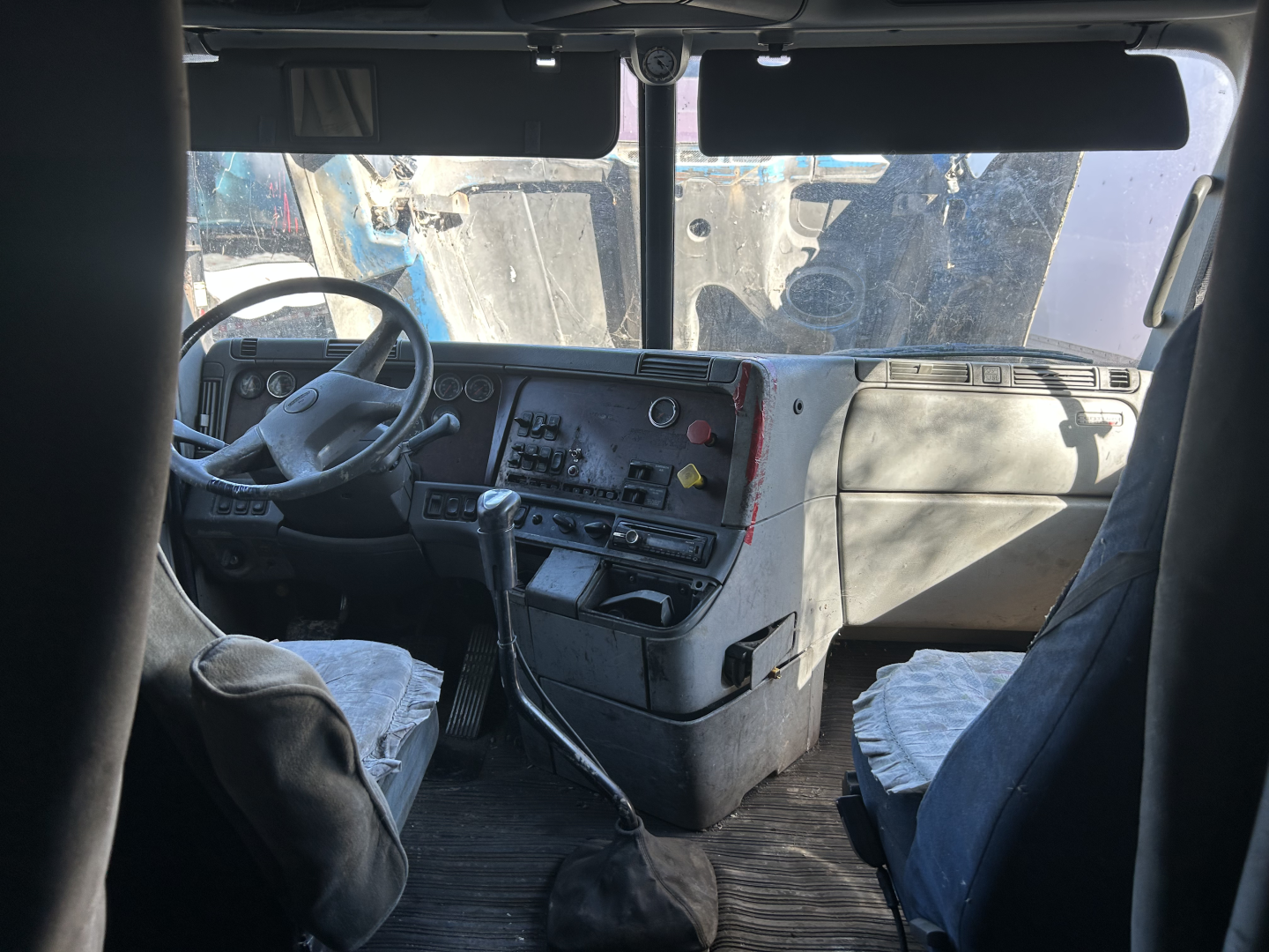 Truck cab interior of a Freightliner FC2 with a left-hand drive dashboard, steering wheel, gear shifter, and two front seats visible.