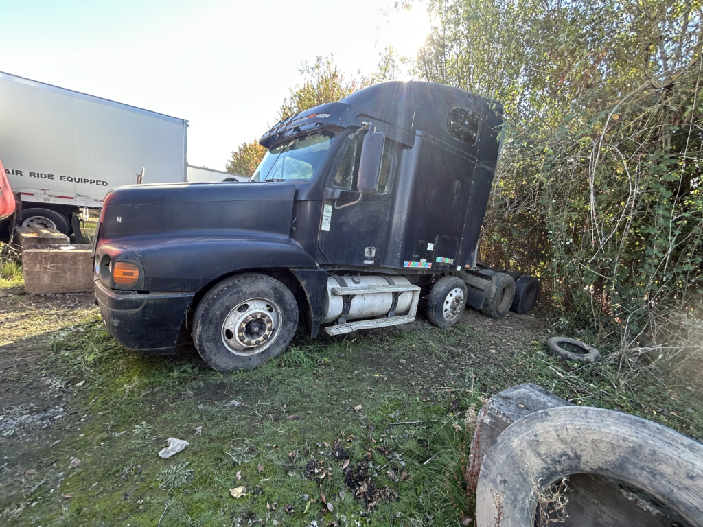 Freightliner conventional Sleeper truck in field with dirt yard and missing front wheel; dark blue cab, large side fuel tanks, dual rear wheels, trees in background.