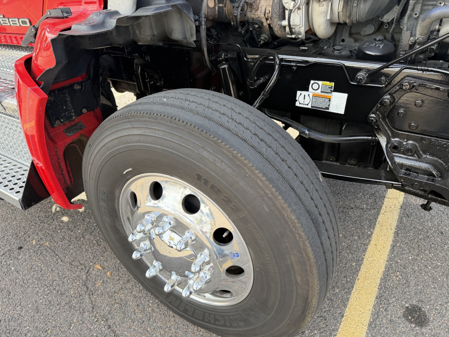 Kenworth T880 front wheel and front suspension area showing large dual rear tires and wheel hub, engine bay with labels and components visible.
