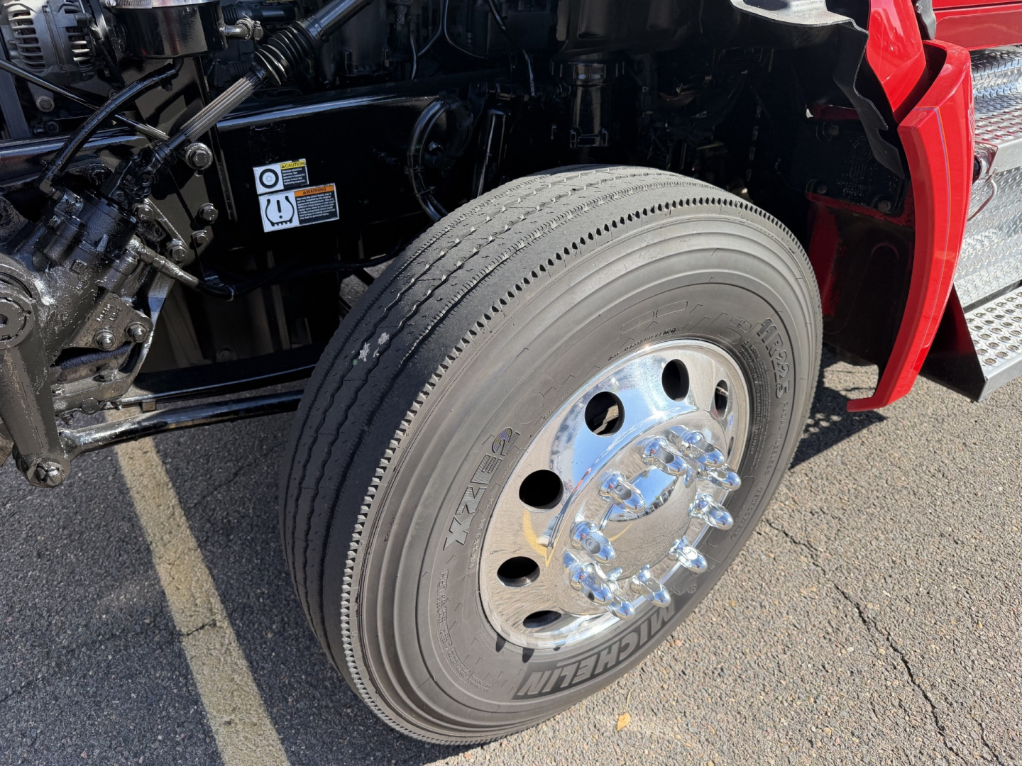 Kenworth T880 front wheel and front suspension visible in a truck yard with chrome lug nuts and steering components.