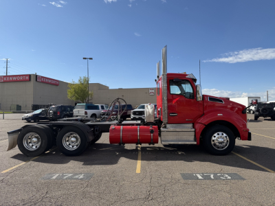 Kenworth T880 tractor on lot with red cab and dual rear axles, side view, with fuel tanks and exhaust stacks visible.