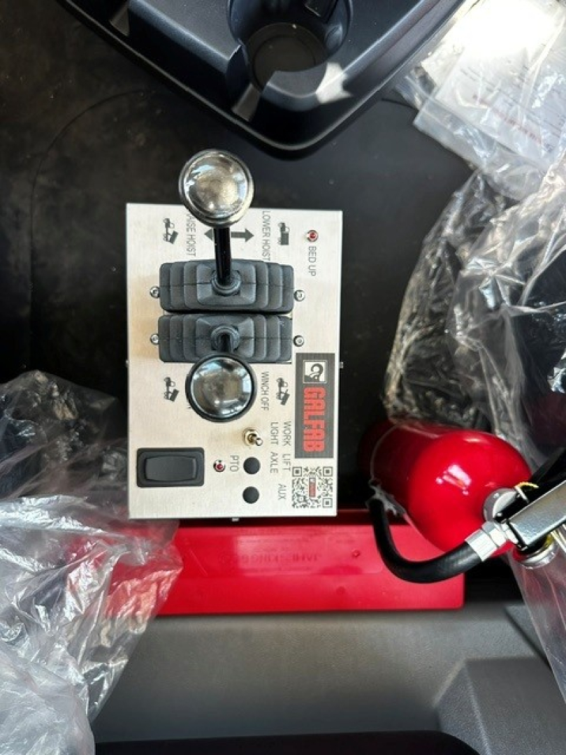 Control panel with two joystick levers and toggle switch, labeled panels around, in a Mack GR64B roll-off truck cab.
