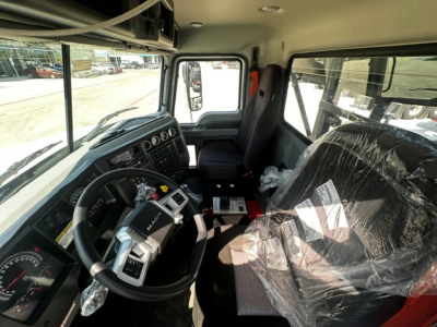 Cab interior of a new 2027 Mack GR64B roll-off truck showing steering wheel, dashboard, driver seat, and plastic-wrapped passenger seat.