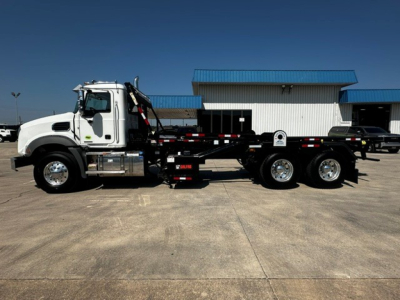 Side view of a white 2027 Mack GR64B roll-off truck with a hydraulic hooklift and long chassis, showing a chrome exhaust, chassis, and dual rear axles.