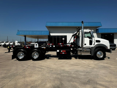Side view of a white Mack GR64B roll-off truck with a black chassis and hydraulic arm, parked in front of a building.