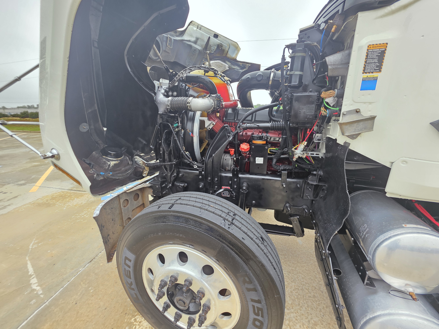Truck engine bay of a 2020 Mack Pinnacle 64T showing engine components, front wheel, and exposed chassis.