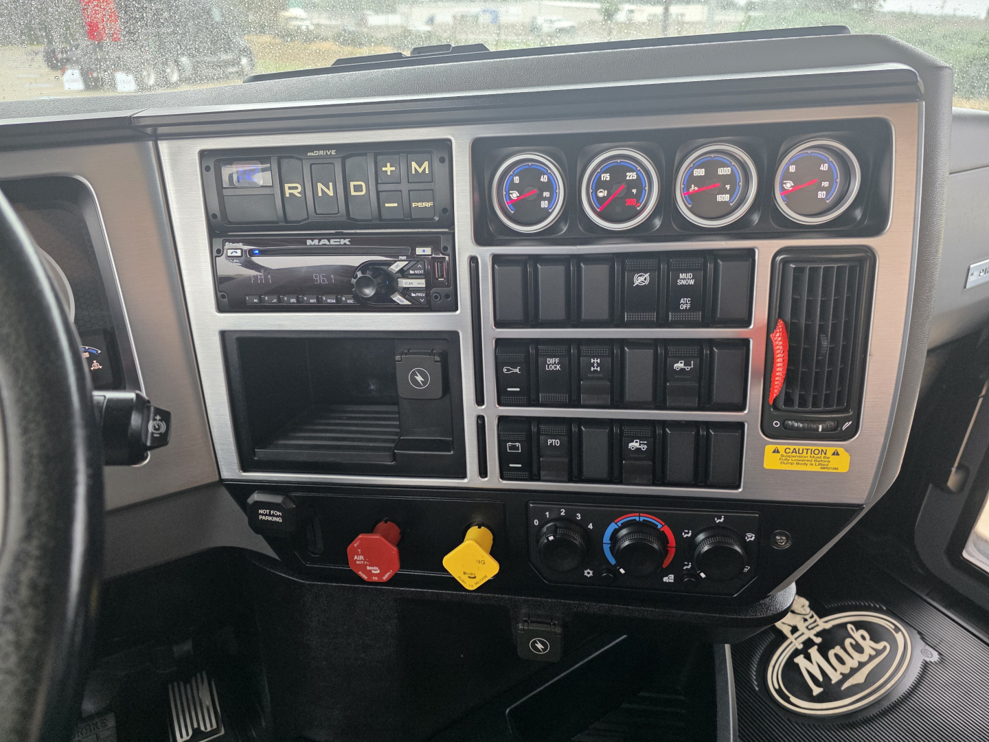 Dashboard of 2020 Mack Pinnacle 64T showing gear selector indicators R-N-D, three round gauges, climate controls, auxiliary switches, ignition, and steering wheel; dash detail with red and yellow knobs.