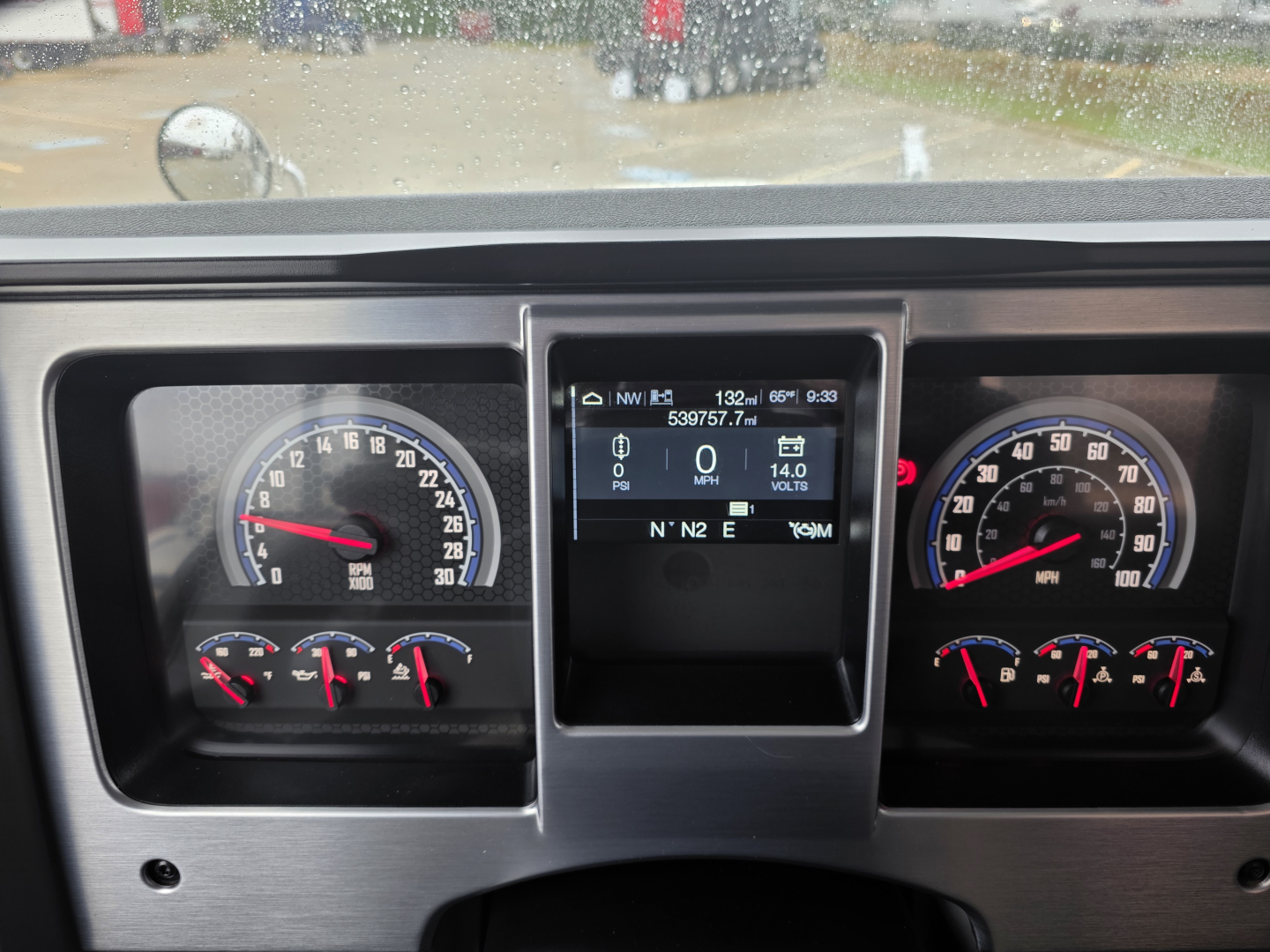 Dashboard of a Mack Pinnacle 64T showing instrument gauges and a central display. Left: speedometer with tachometer and smaller gauges. Right: additional gauges. Center: digital display panel.