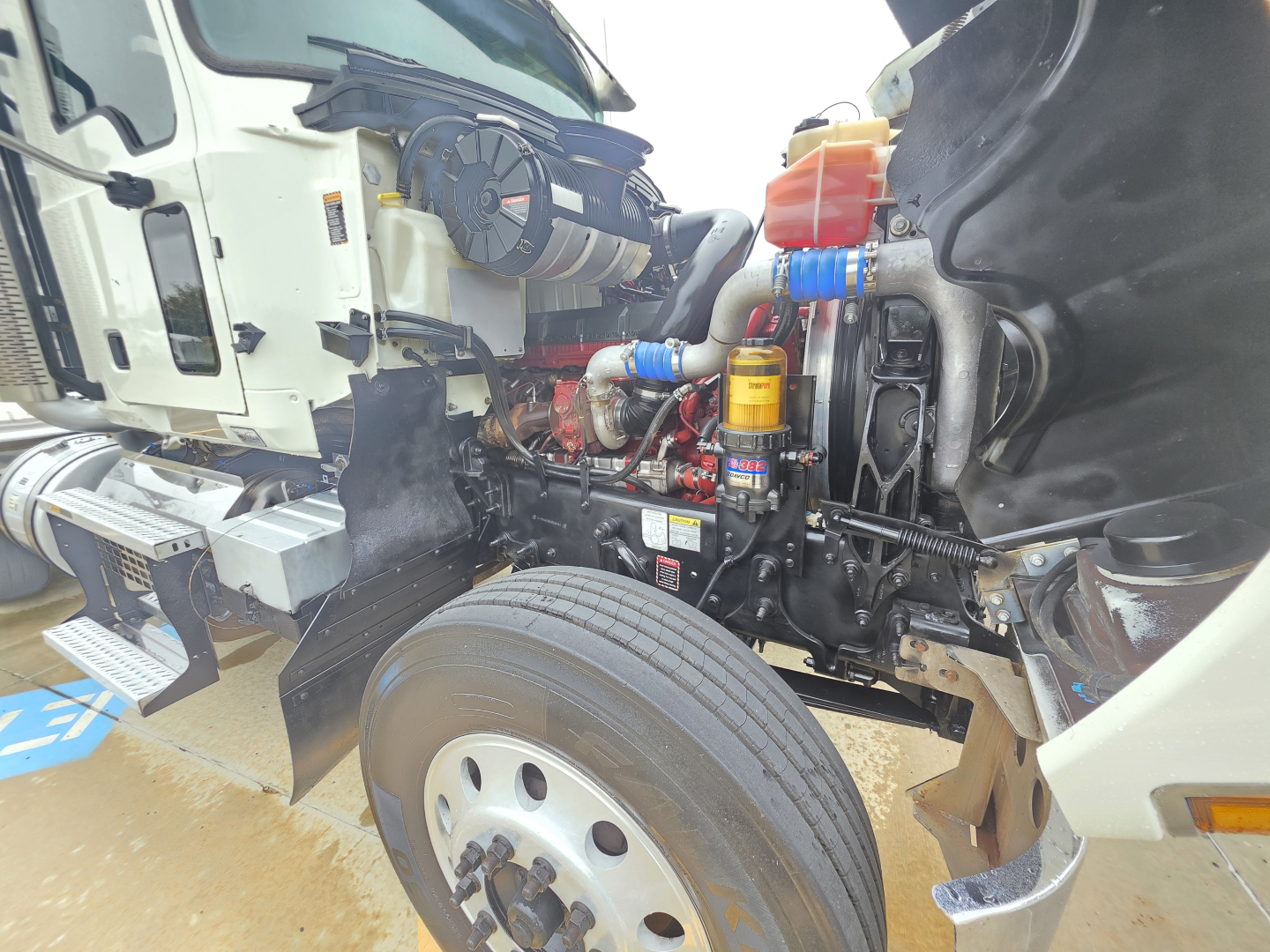 Mack Pinnacle 64T truck front left showing engine bay components and wheel with blue hoses and orange coolant reservoir.