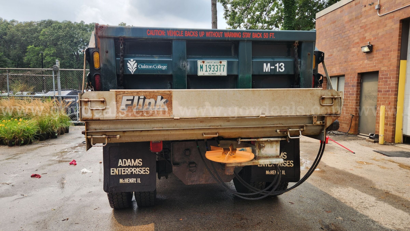 Back view of a blue/green dump truck bed with Shelley/Fuck branding and a spare wheel, license plate, and hydraulic hoses visible.