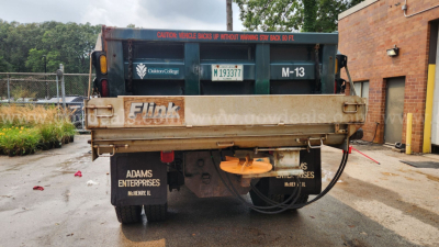 Back view of a blue/green dump truck bed with Shelley/Fuck branding and a spare wheel, license plate, and hydraulic hoses visible.