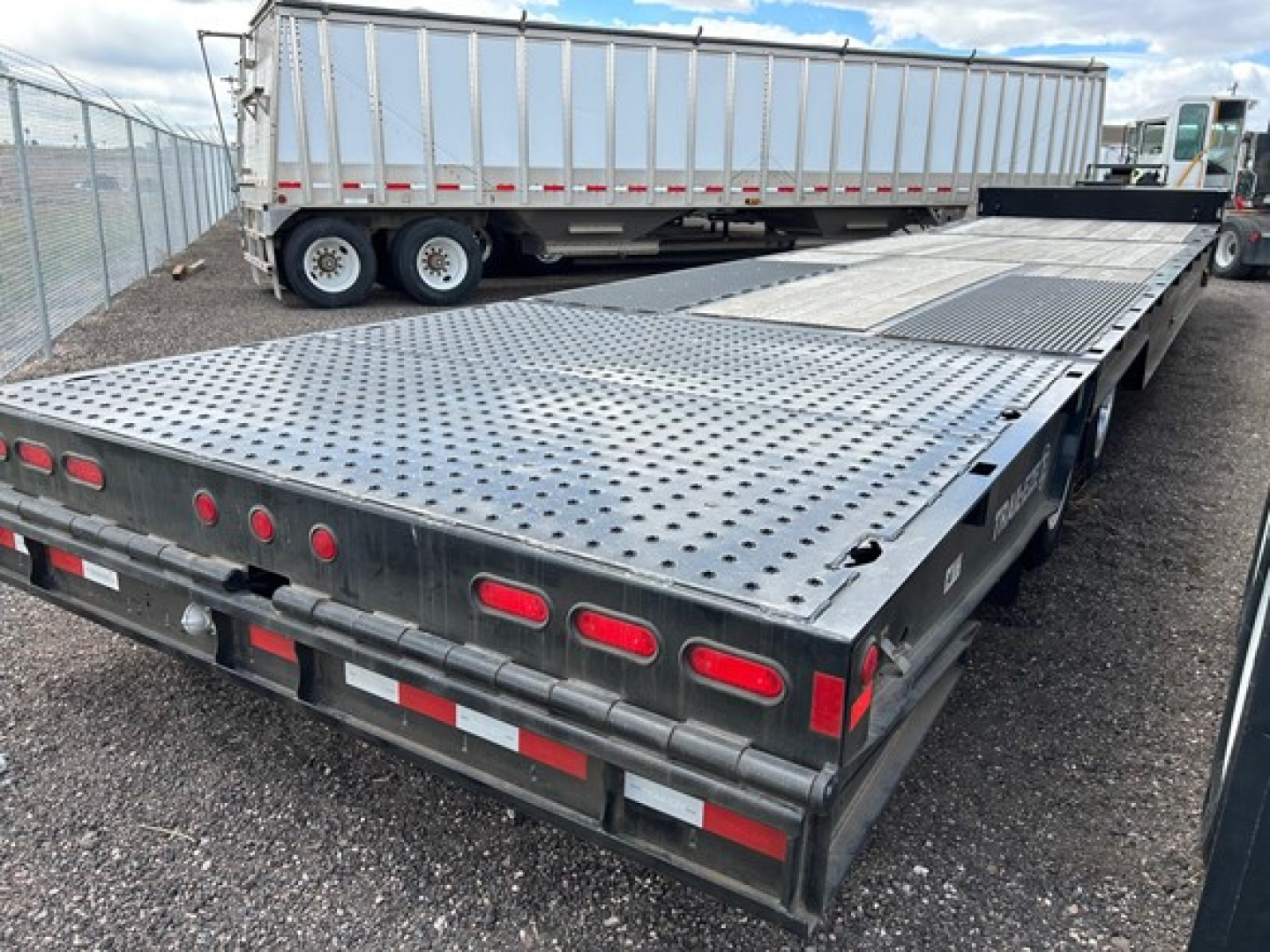 Flatbed trailer with perforated steel deck and rear LED lights in view; accompanying semi-trailer with tall silver trailer in background.