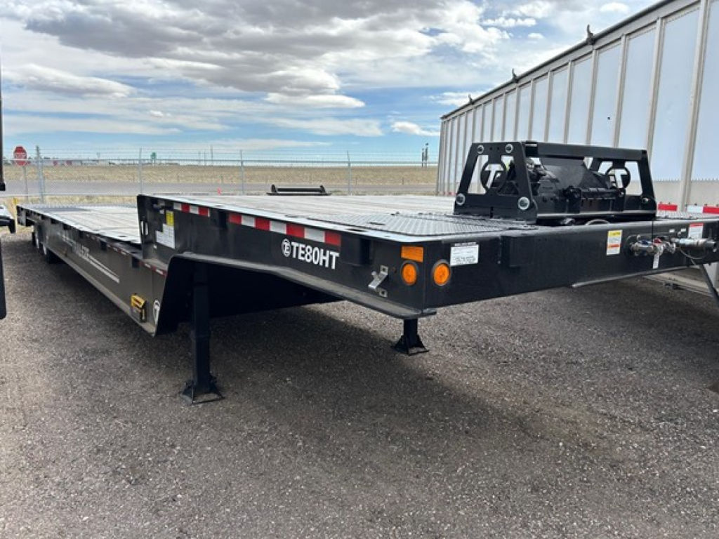 Trail-Eze TE80HT flatbed trailer showing TE80HT branding, front hitch area, black steel frame, orange reflectors, and support legs on a gravel lot.